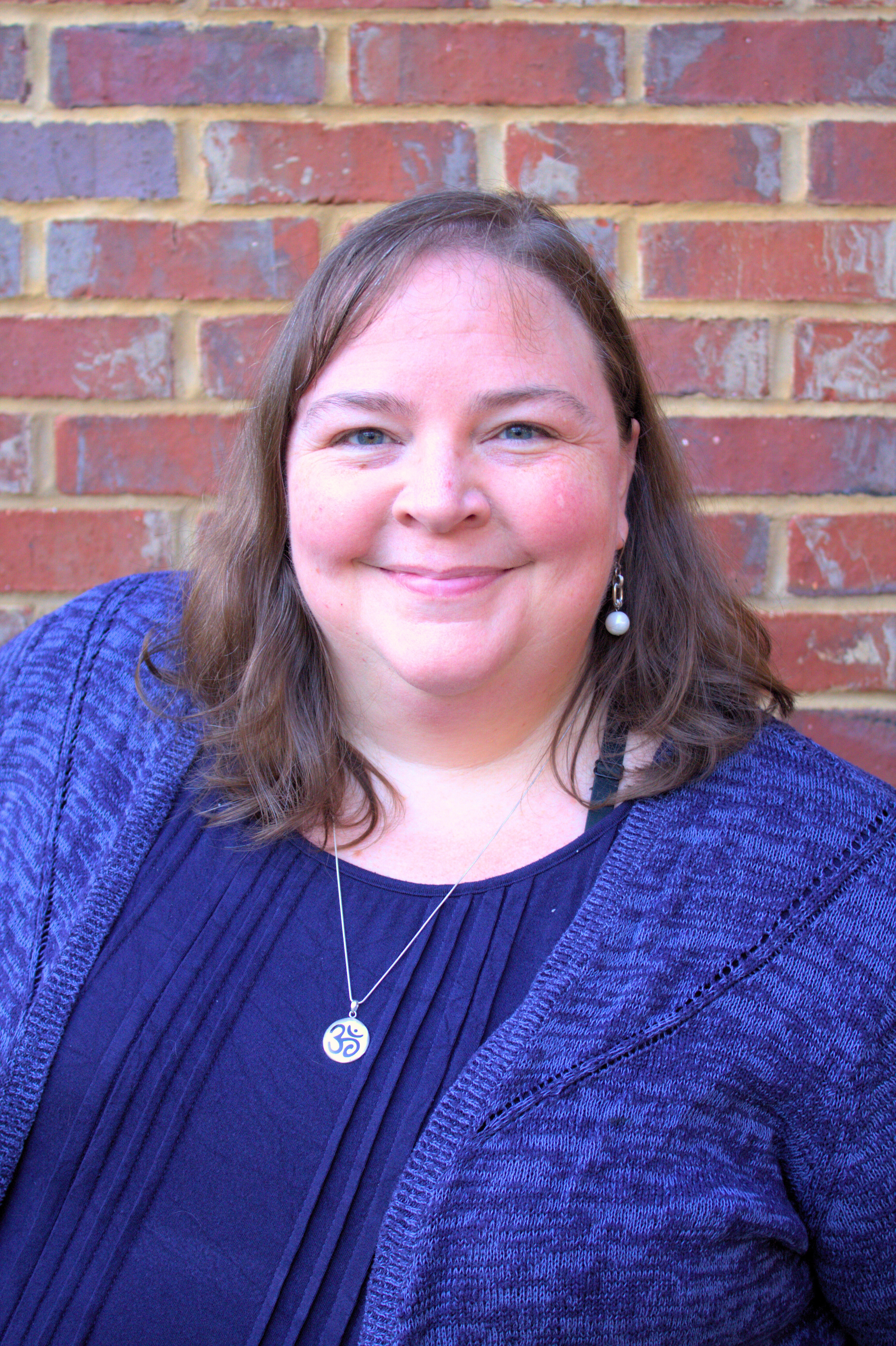 Liz Norell is a White woman in a blue shirt and blue cardigan. She stands in front of a red brick wall and wears a silver necklace that says "ohm" in sanskrit. She has medium-length brown hair and a round face.