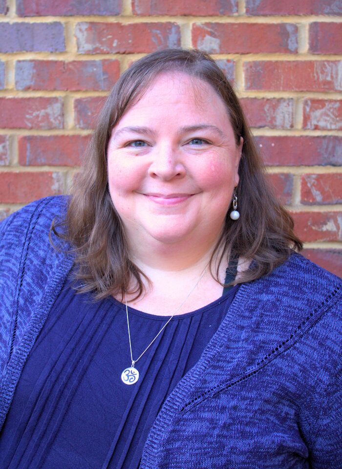 Liz Norell, a white woman with shoulder-length brown hair and a round face, wears a navy shirt, navy/bright blue cardigan, and a silver necklace with the ohm symbol, stands in front of a red brick wall.